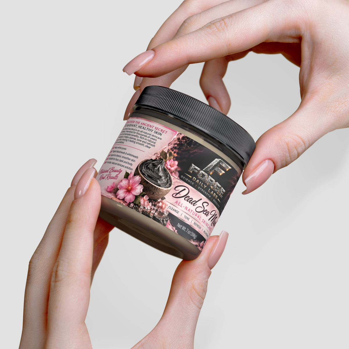 Hands holding a jar of Dead Sea Salt bath salts against a light gray background. The jar has a black lid and a label with pink floral graphics and black text.